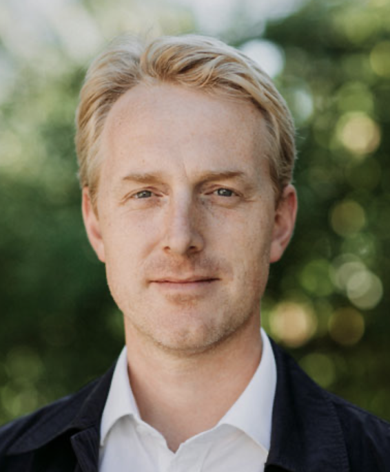 A young man with blonde hair and blue eyes, wearing a white collared shirt and dark blazer, outdoors with blurred greenery in the background.