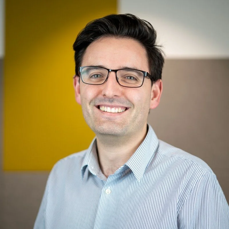 Portrait of a man with dark hair, glasses, and a striped shirt, smiling in front of a colorful background.
