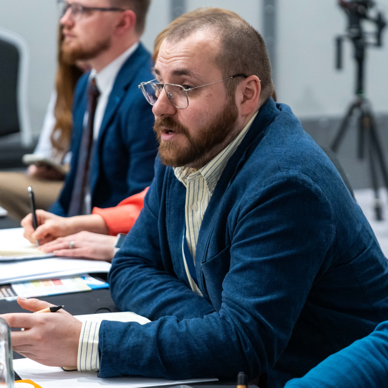 A man with glasses, a beard, and short hair wearing a blazer and striped shirt, sitting at a conference table during a meeting.