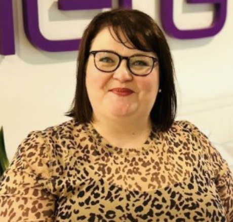 A woman with shoulder-length dark hair, wearing glasses and a leopard print top, standing indoors with a purple sign in the background.