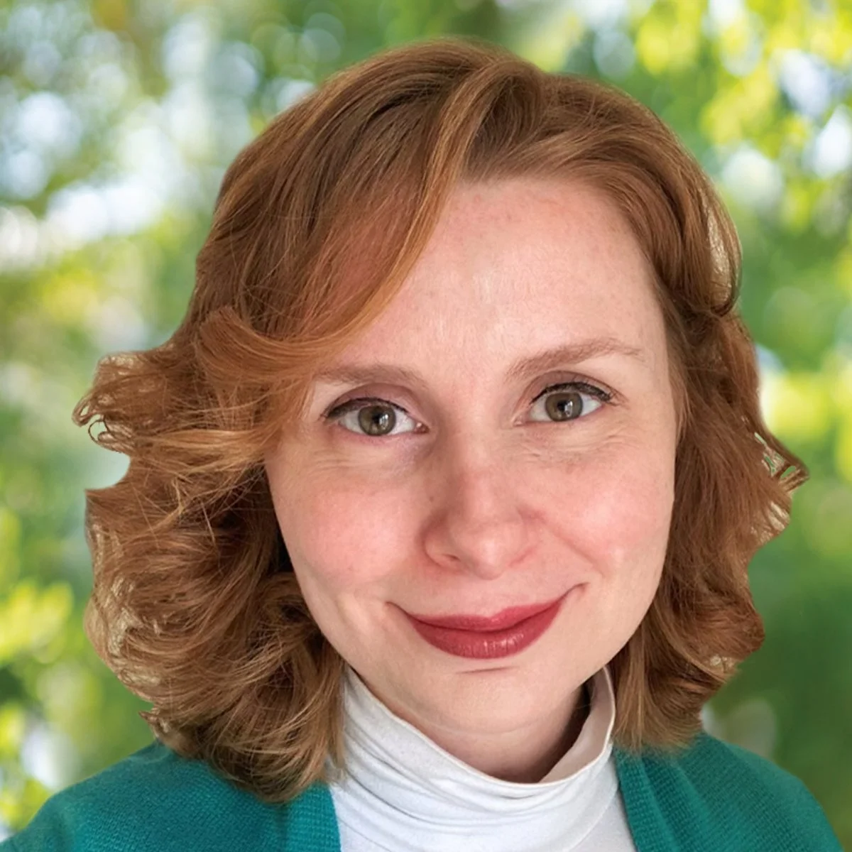 Close-up of a woman with red hair and fair skin smiling, outdoors with blurred green trees in the background.