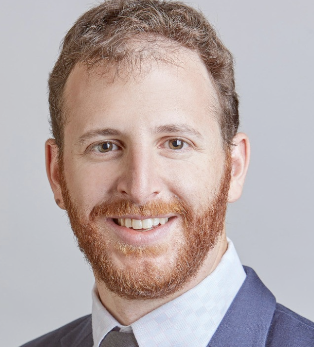 A smiling man with a red beard and short, curly hair, wearing a business suit jacket, shirt, and tie, posing against a plain background.