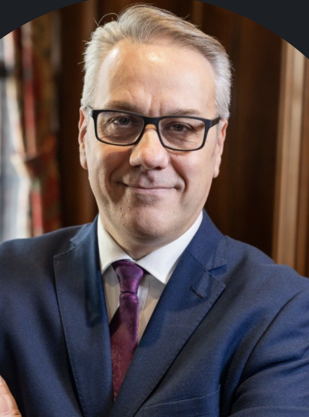 A middle-aged man wearing a suit and tie, glasses, smiling, with a bookshelf in the background.