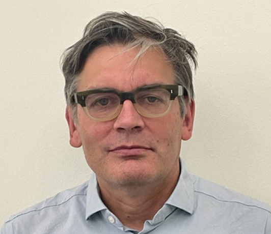 A middle-aged man with gray hair and glasses wearing a light blue button-up shirt, looking directly at the camera against a plain background.