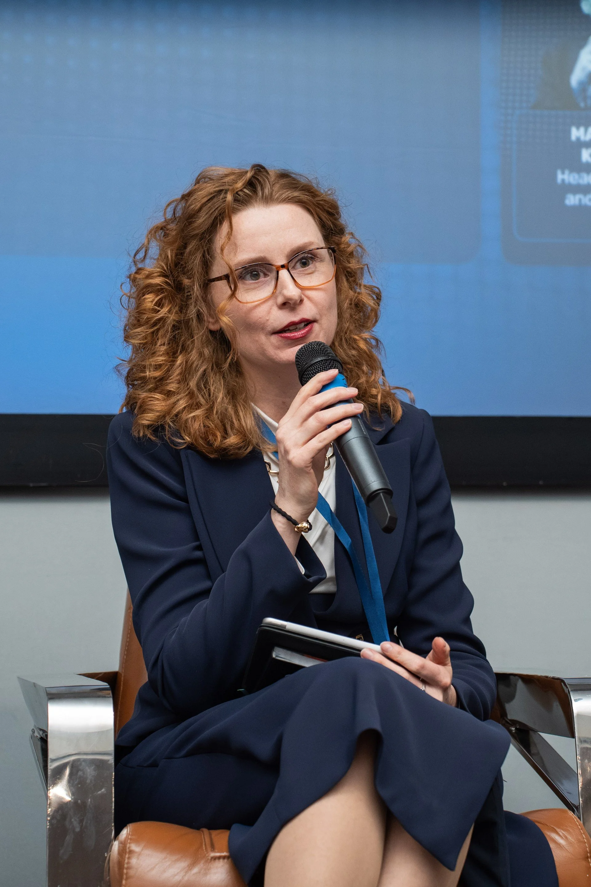 A woman with curly red hair and glasses sitting on a leather chair, holding a microphone and a notepad, speaking at an event.