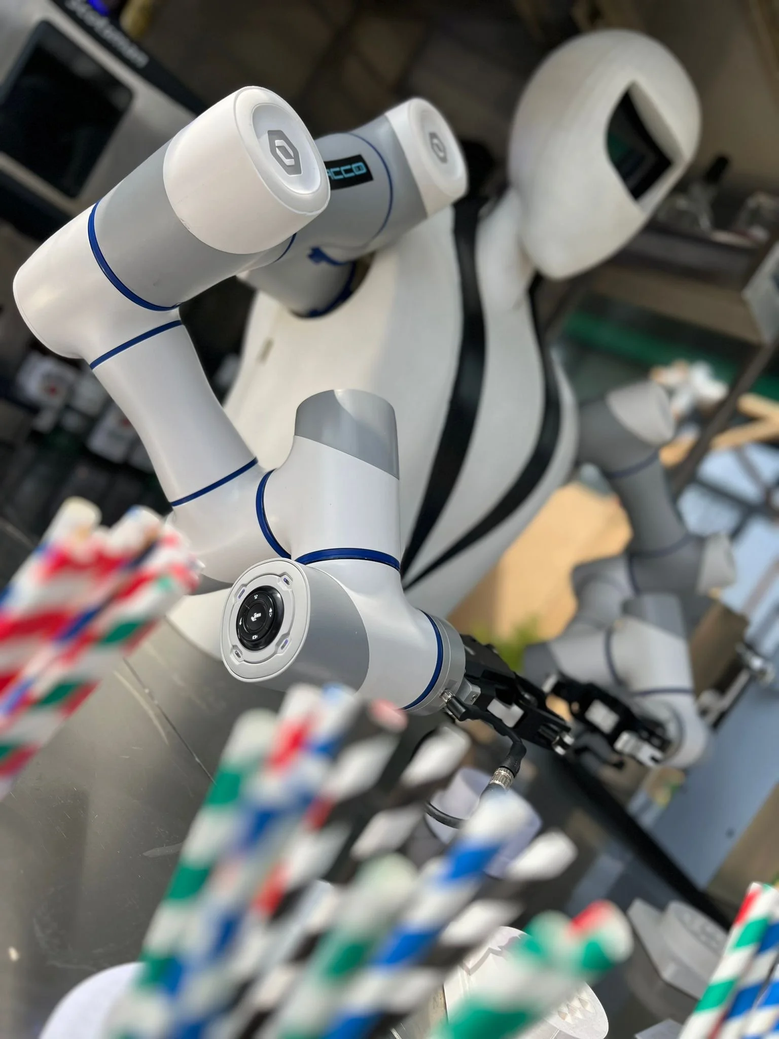 Robot humanoïde blanc et gris avec des articulations en bleu, debout dans un atelier, avec des pailles colorées floues au premier plan.