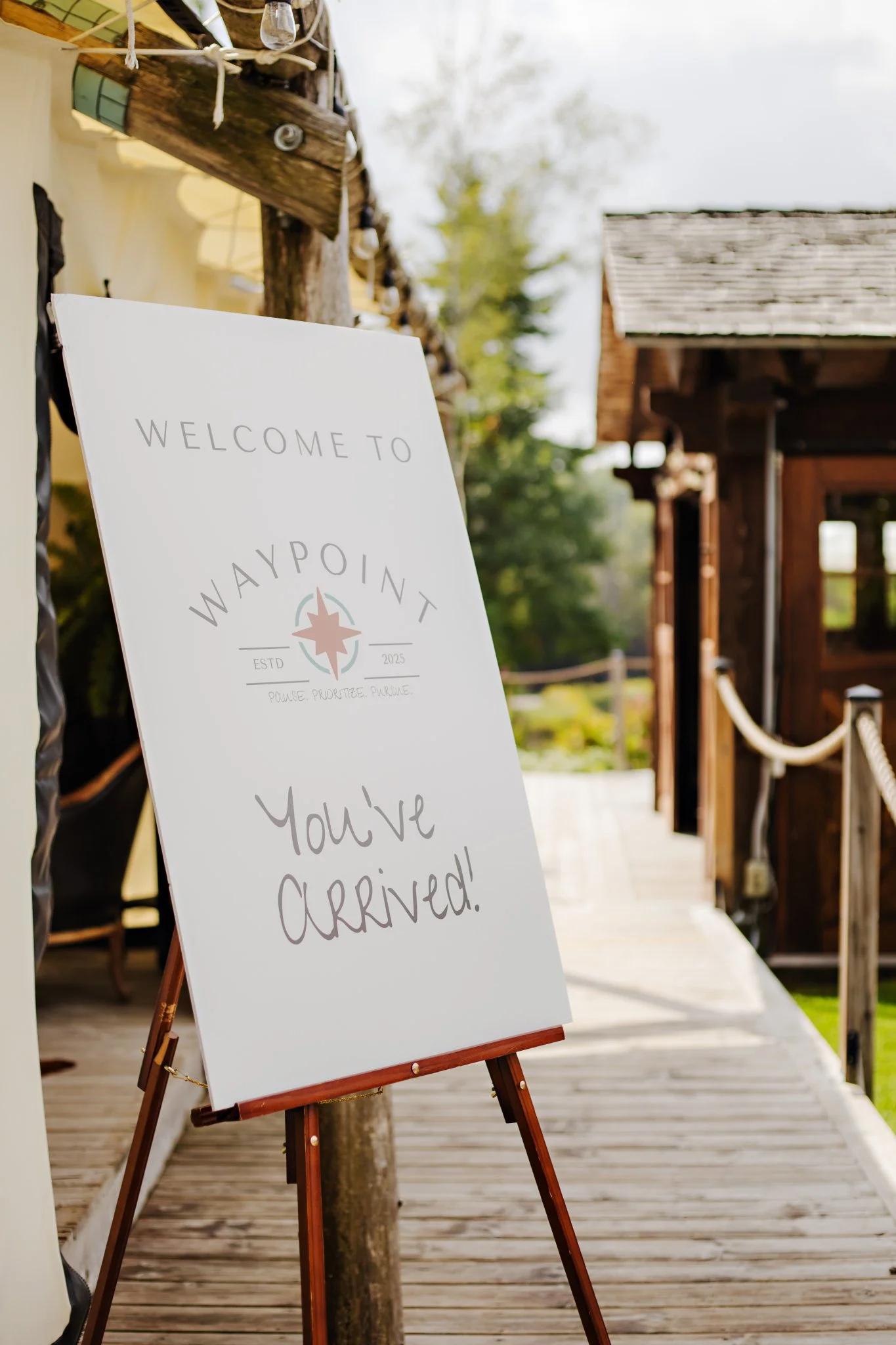 Welcome sign on an easel that says, 'You’ve arrived!', at an outdoor venue with wooden buildings and a pathway, possibly for an event or gathering.