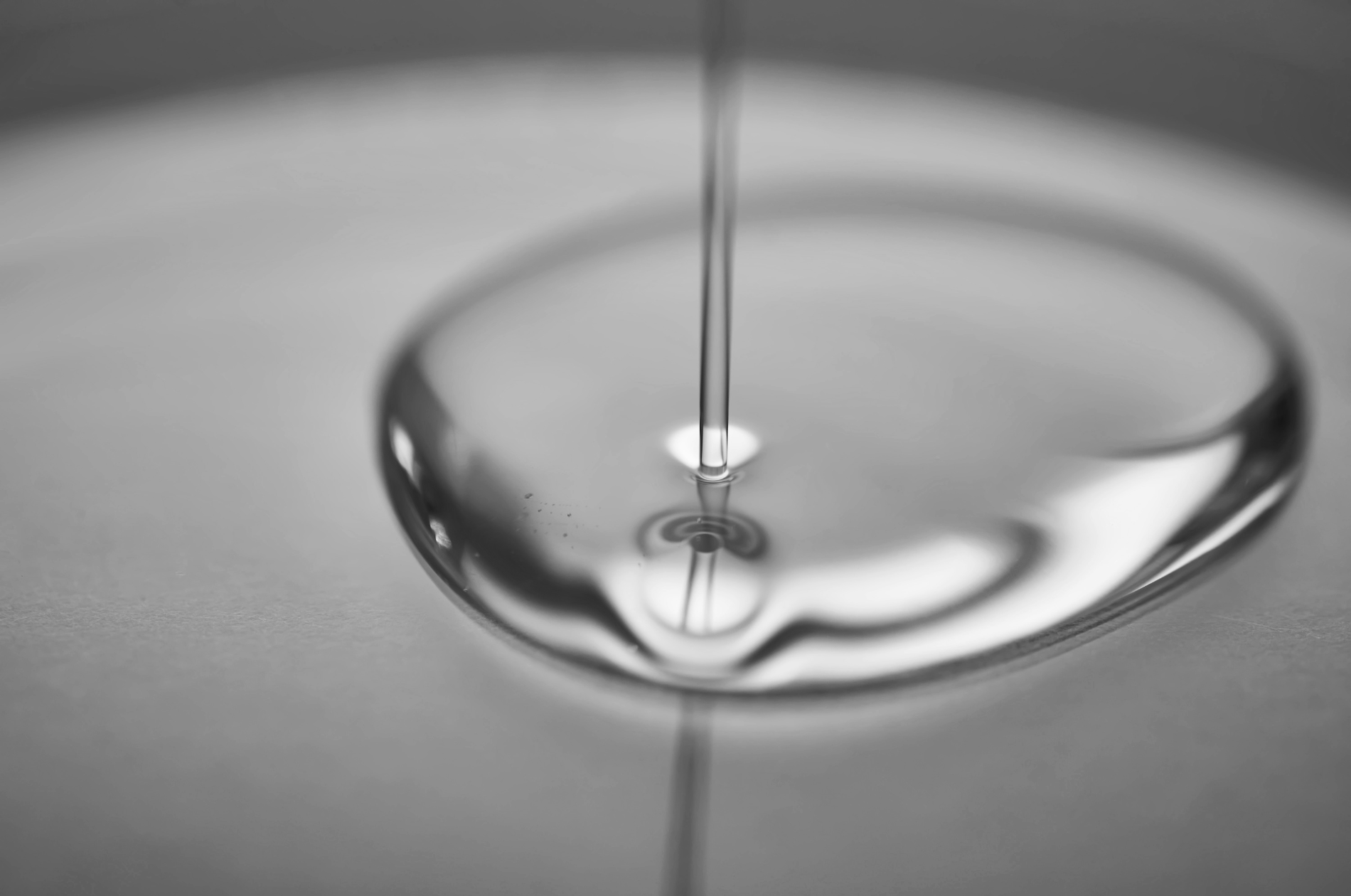 Close-up of a thin stream of water falling into a shallow pool, creating ripples and a small splash in black and white.