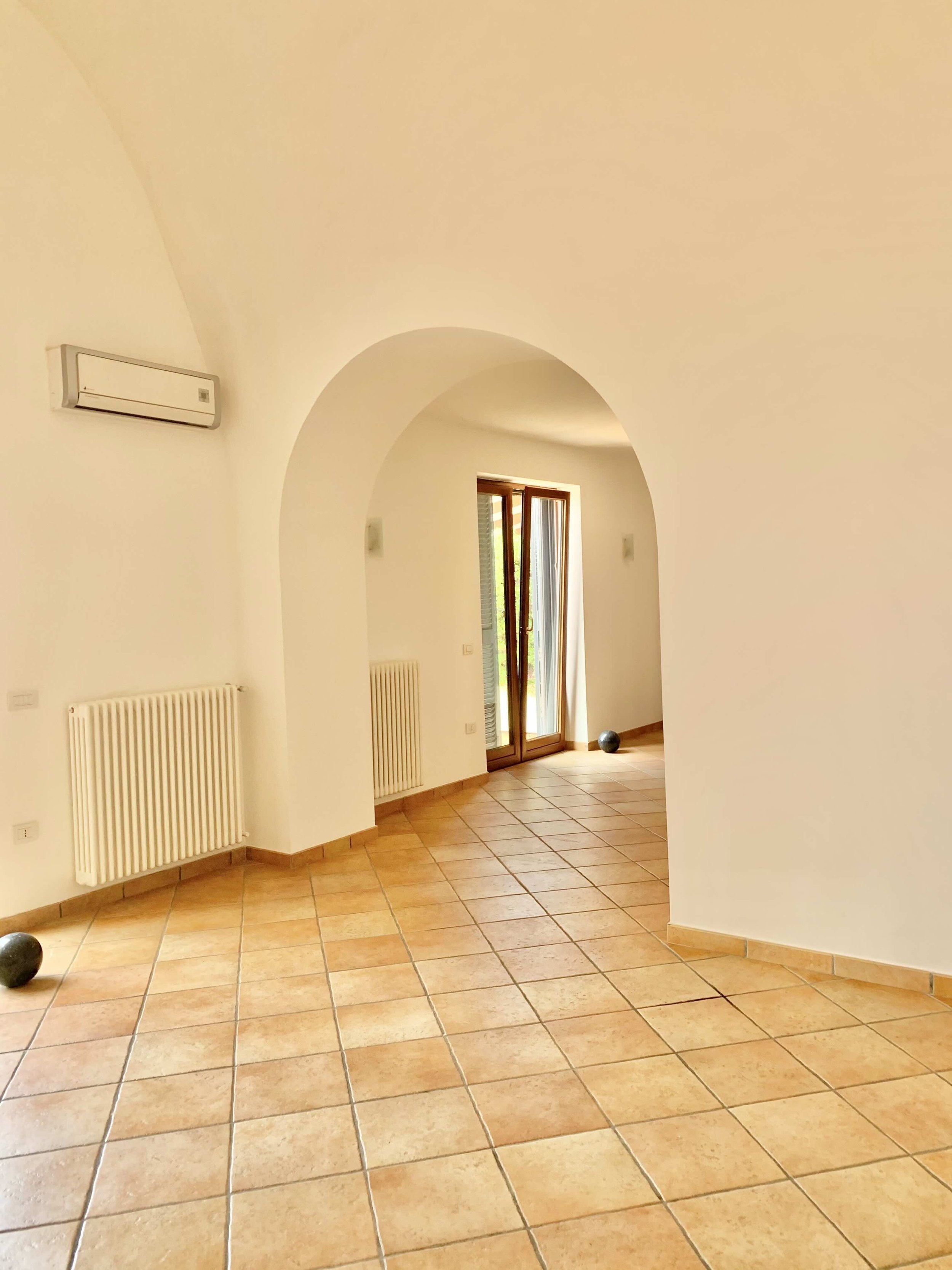 Empty room with arched doorway, beige tiled floor, white walls, air conditioning unit, radiator, and wooden framed window.