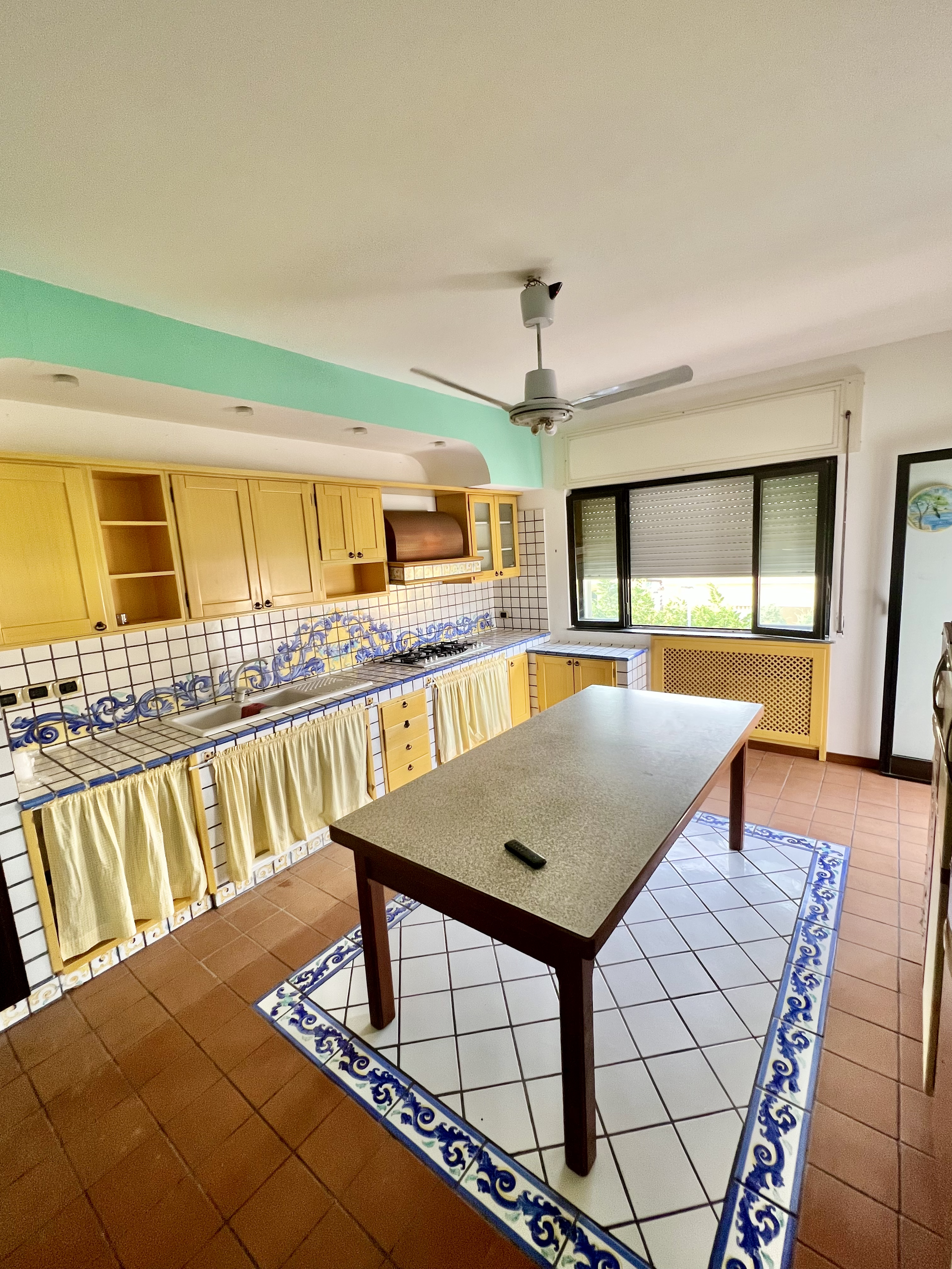 Traditional kitchen with wooden cabinets, tiled countertops, decorative tiles, a central table, ceiling fan, and large window with a view.