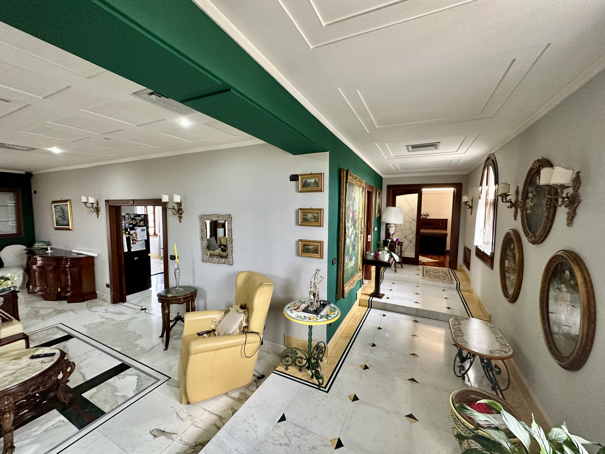 Elegant interior of a living area with marble floors, a yellow armchair, ornate wooden furniture, decorative wall sconces, framed paintings, and mirrors. A hallway leads to another room, featuring plants and accent tables.