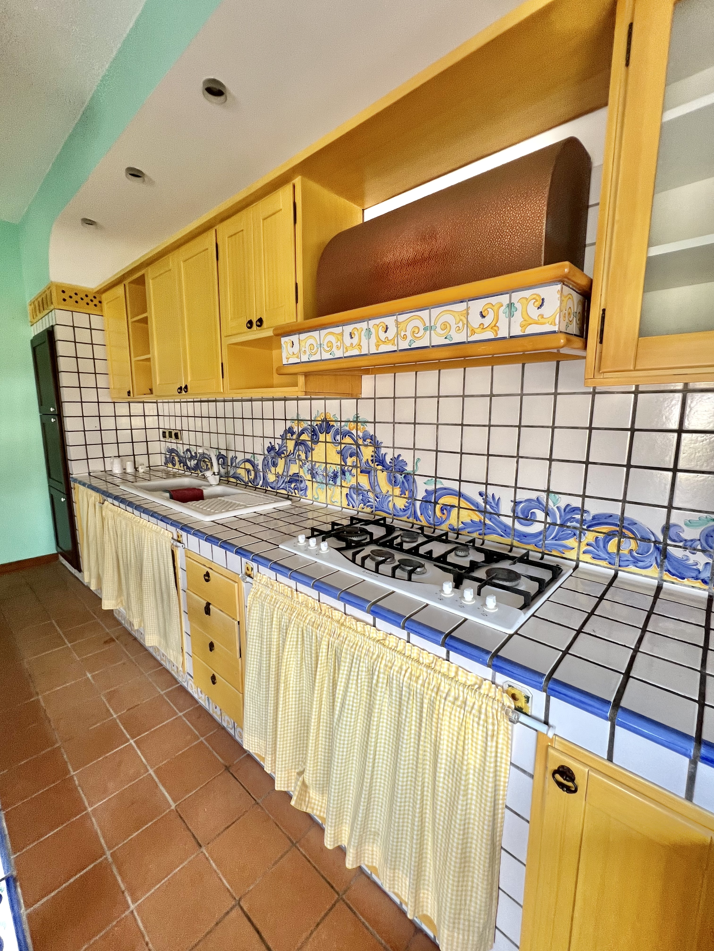 Bright kitchen with yellow wooden cabinets, decorative tiles, gas stove, and checkered curtains beneath the counter.
