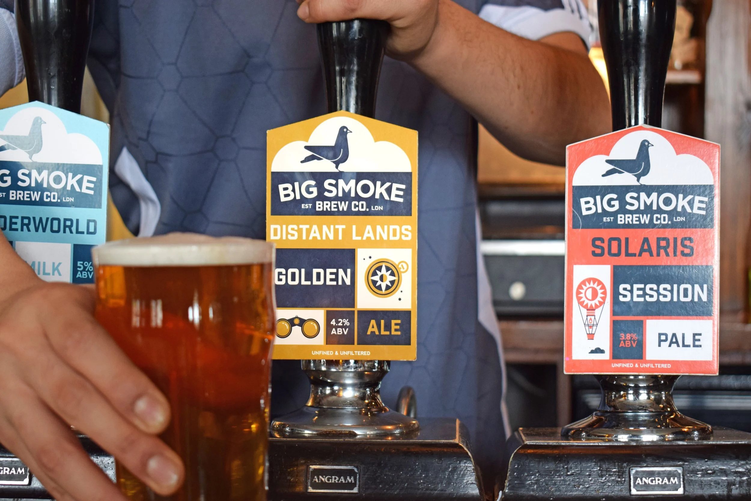 A person holding a glass of amber beer with a foamy head near two taps of Big Smoke Brew Co. beers. The left tap offers 'Distant Lands Golden Ale' with a 4.2% ABV, and the right tap offers 'Solaris Session Pale' with a 3.8% ABV.