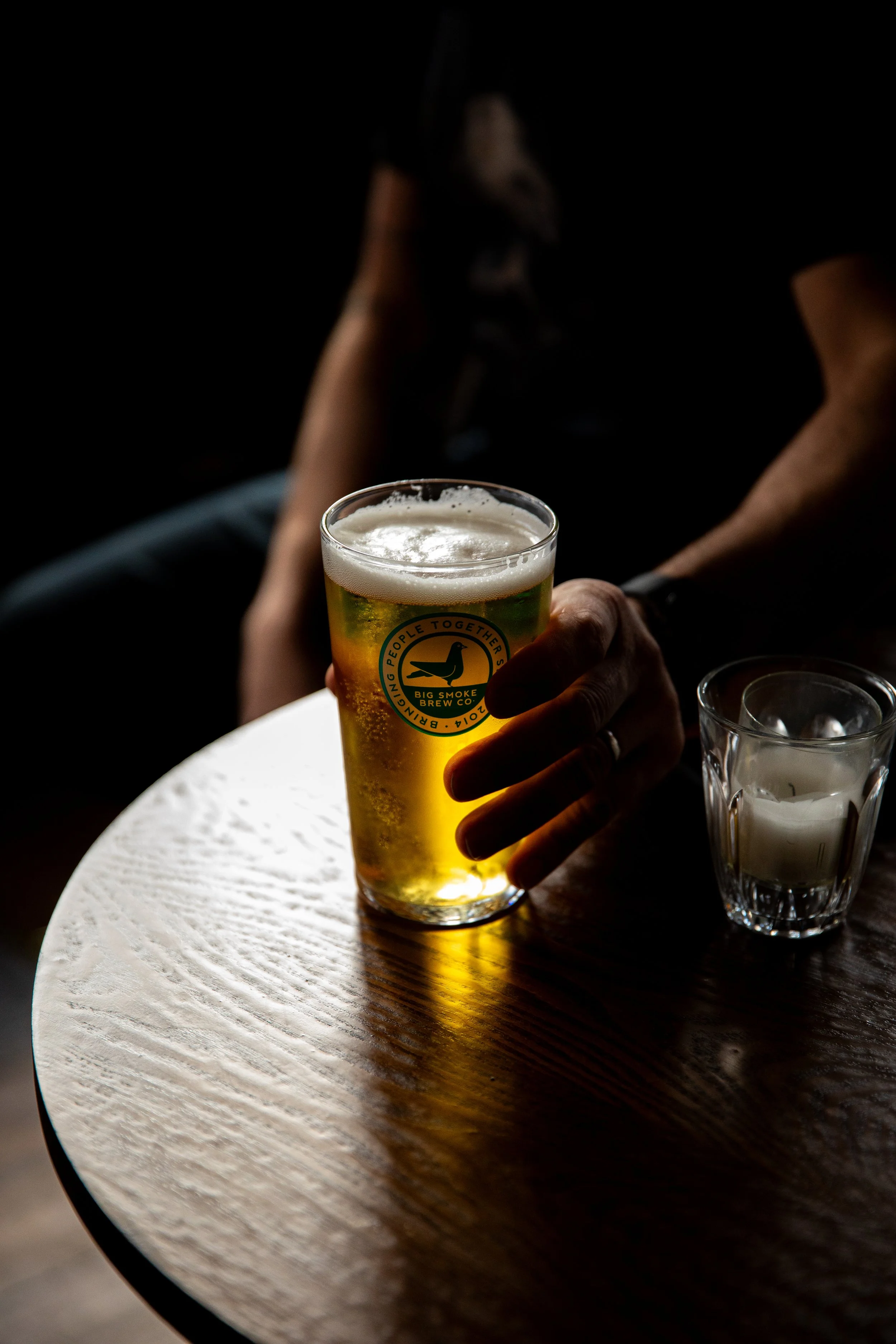 A person holding a glass of beer on a wooden table, with a candle in a glass holder beside it. The background is dark and blurred.