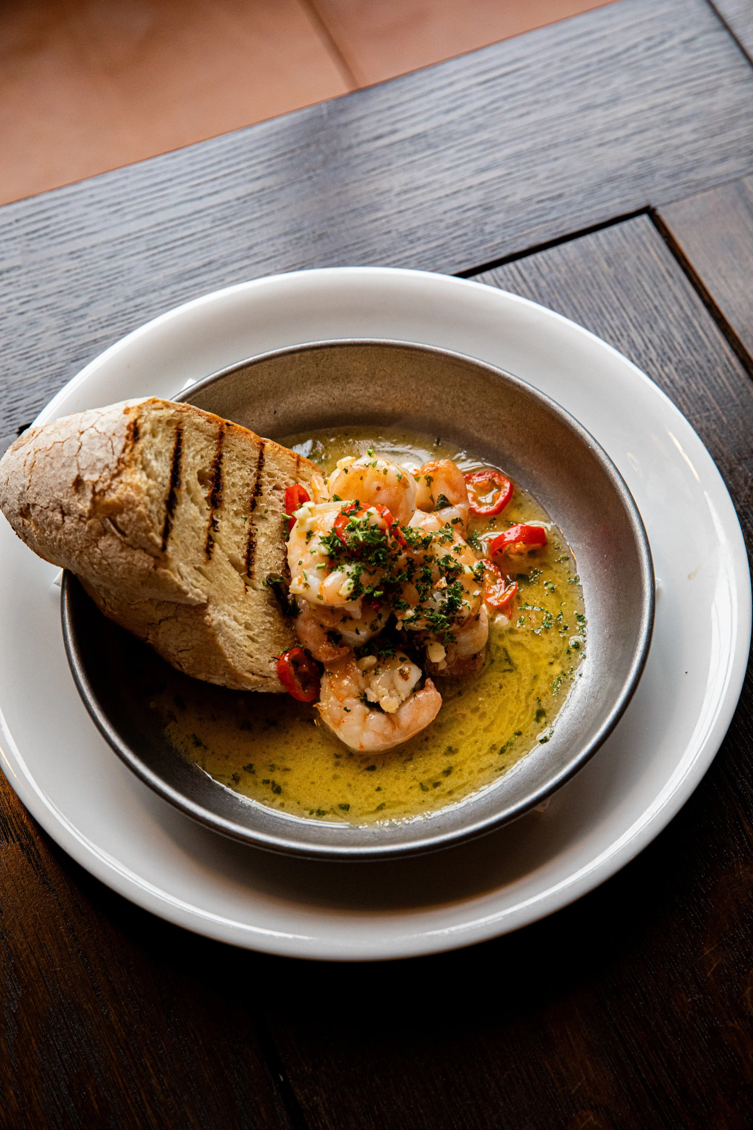 Shrimp served in a bowl with sauce, garnished with herbs, accompanied by a slice of grilled bread, on a white plate.