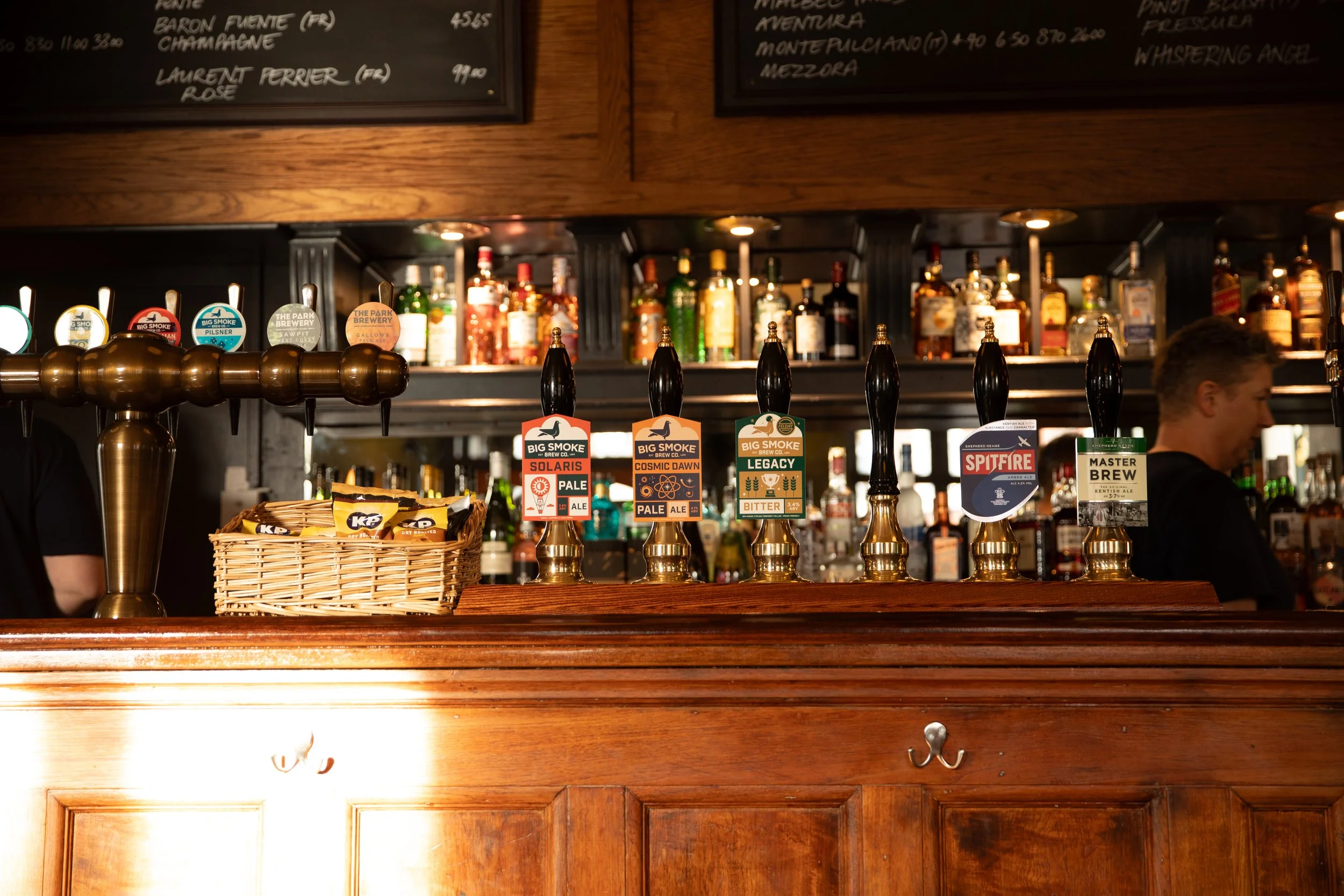 A bar with six beer taps labeled Solaris Pale, Cosmic Dawn Pale Ale, Legacy Bitter, Spitfire, and Master Brew. Behind the bar are shelves with bottles of liquor and a chalkboard menu above. A bartender is partially visible on the right.