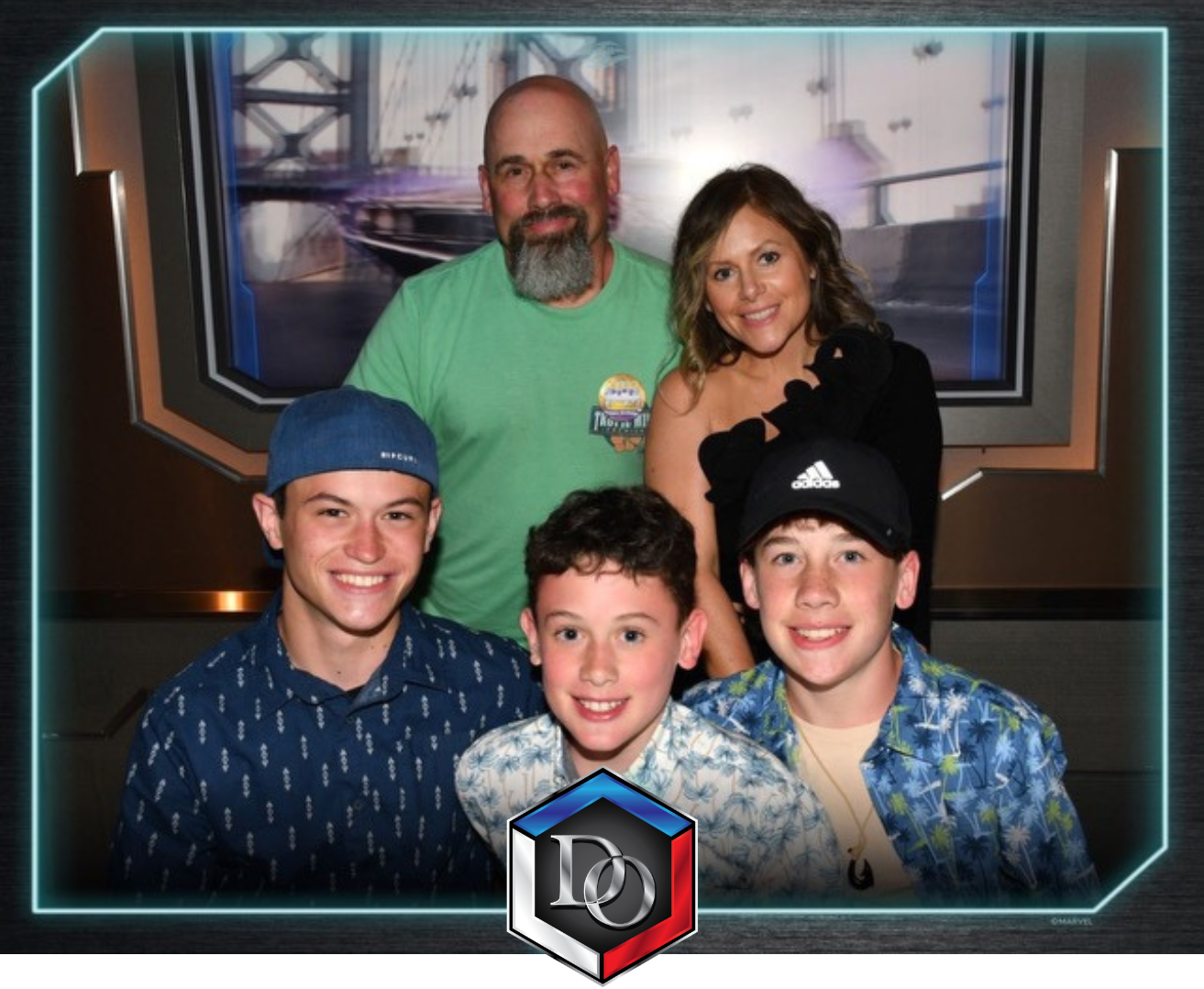 A group photo of two adults and three children smiling indoors, with a futuristic digital border and a Marvel logo at the bottom. The background shows a display with a spaceship or aircraft image.