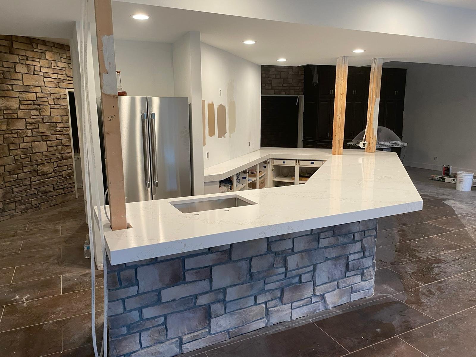 Modern kitchen under renovation with stone accent wall, unfinished white countertops, sink, refrigerator, and partially constructed cabinets.