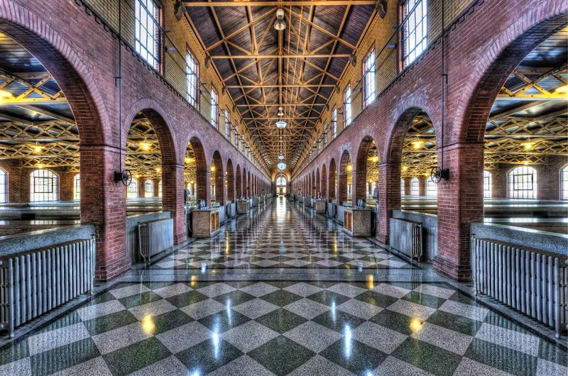 Interior of a historical building with arched brick architecture and checkered floor, featuring large windows and an intricate wooden ceiling.
