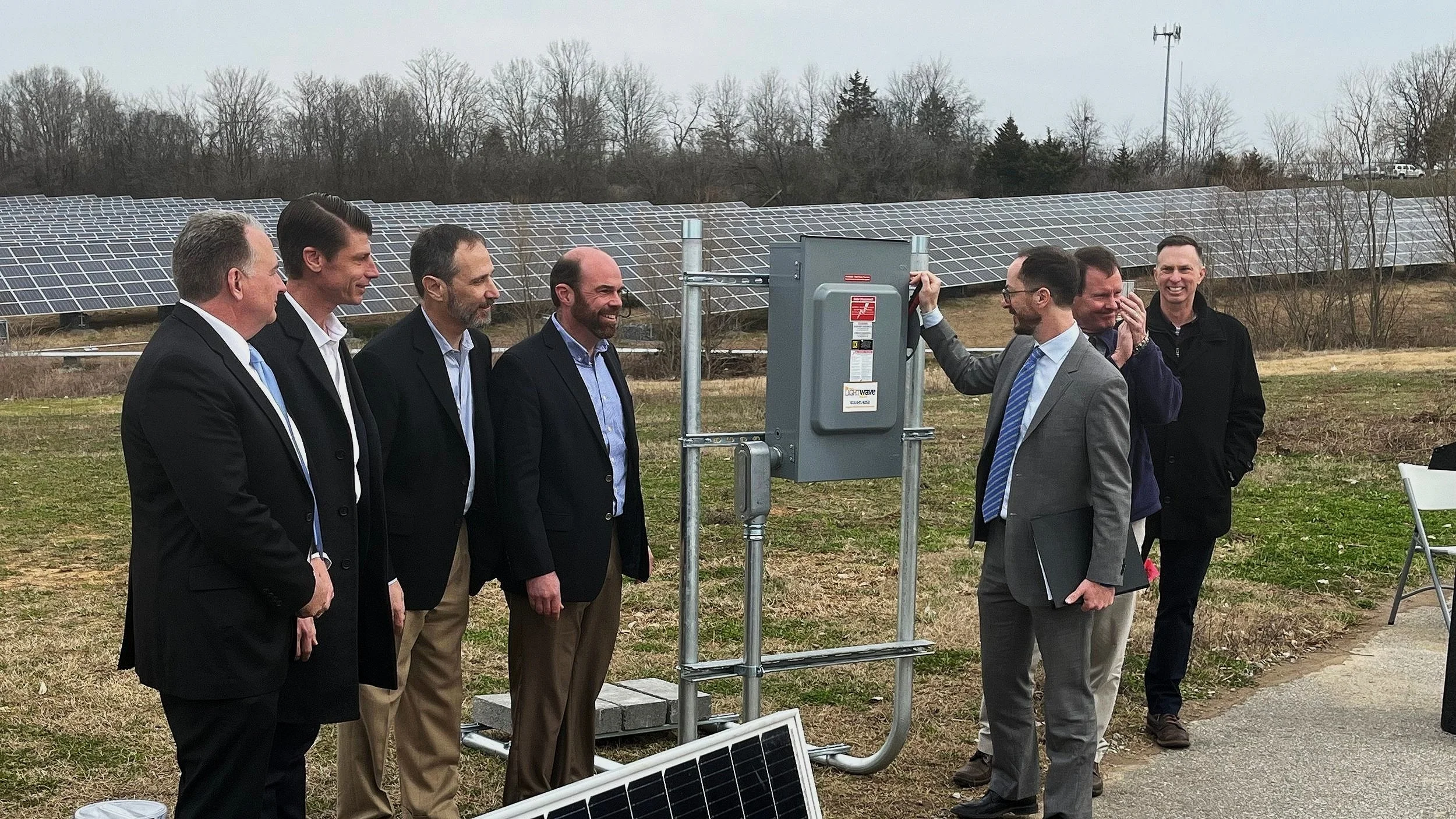 Group of men in suits standing near solar panels at an outdoor installation site.