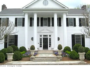 A large white colonial-style house with four columns and a symmetrical design, featuring black shutters and a landscaped garden with shrubs and flowers.