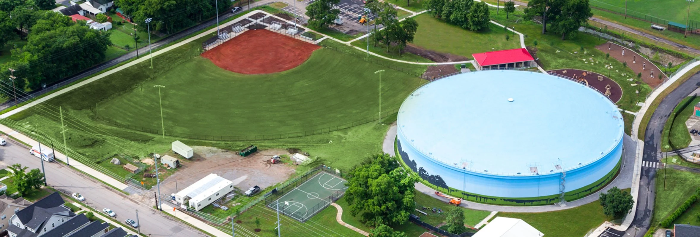 Aerial view of a sports facility with a large circular blue building, a baseball field with red dirt, a small basketball court, and adjacent green spaces and parking areas.