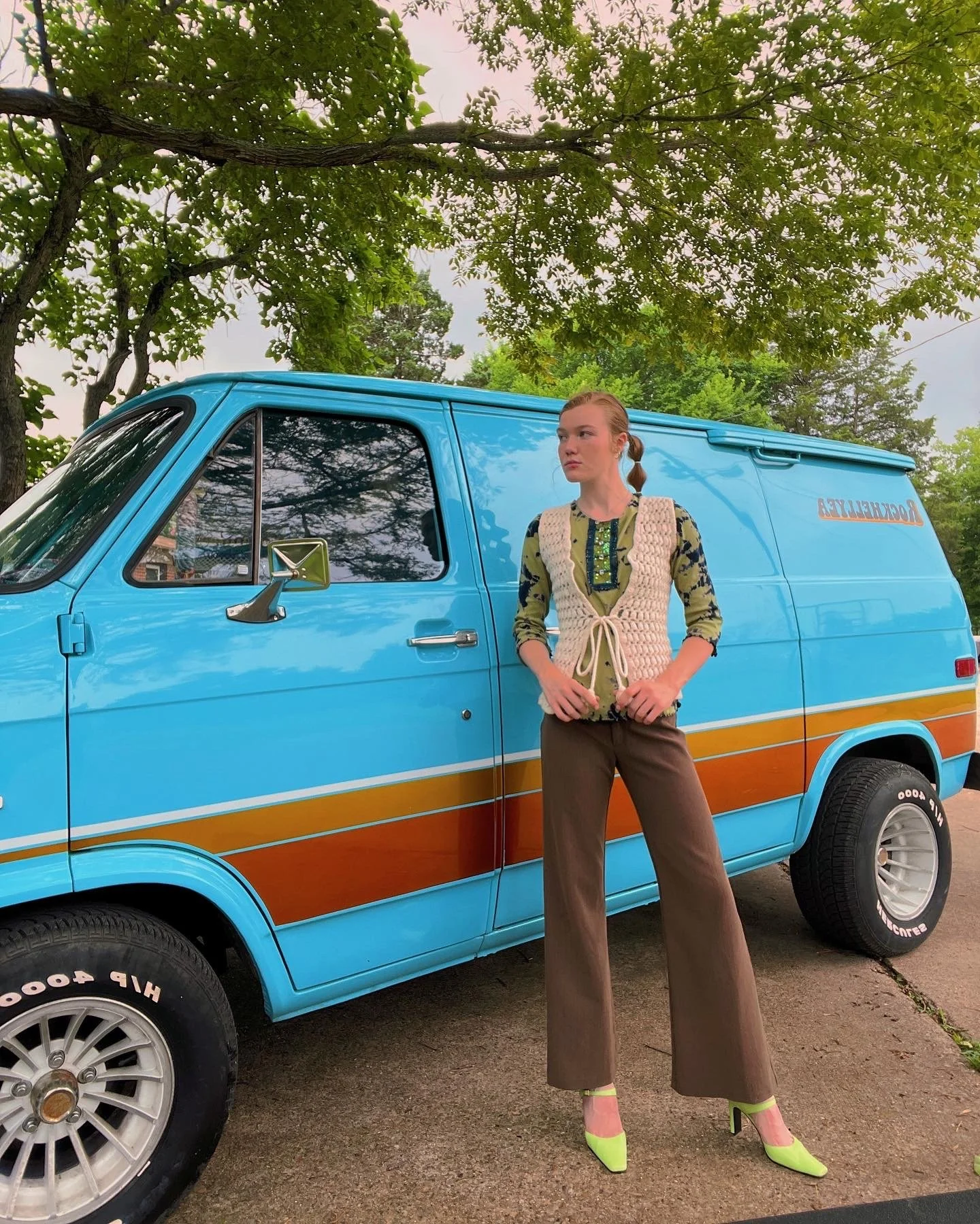 A woman standing next to a vintage blue van with orange and yellow stripe details, under the branches of a large tree.