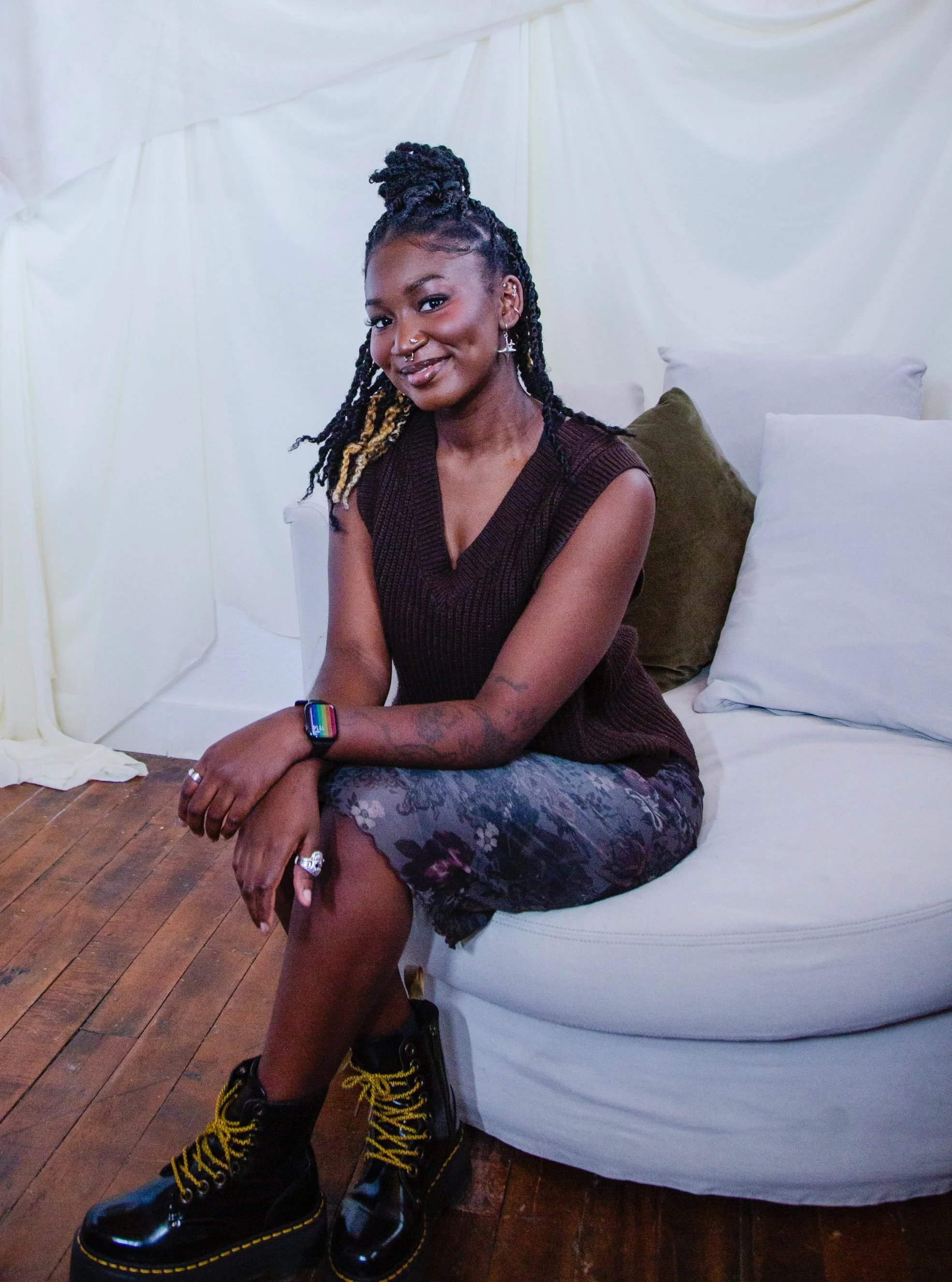 A woman with braided hair and tattoos, sitting on a white sofa, smiling, wearing a dark sleeveless top, patterned skirt, and black boots with yellow laces.