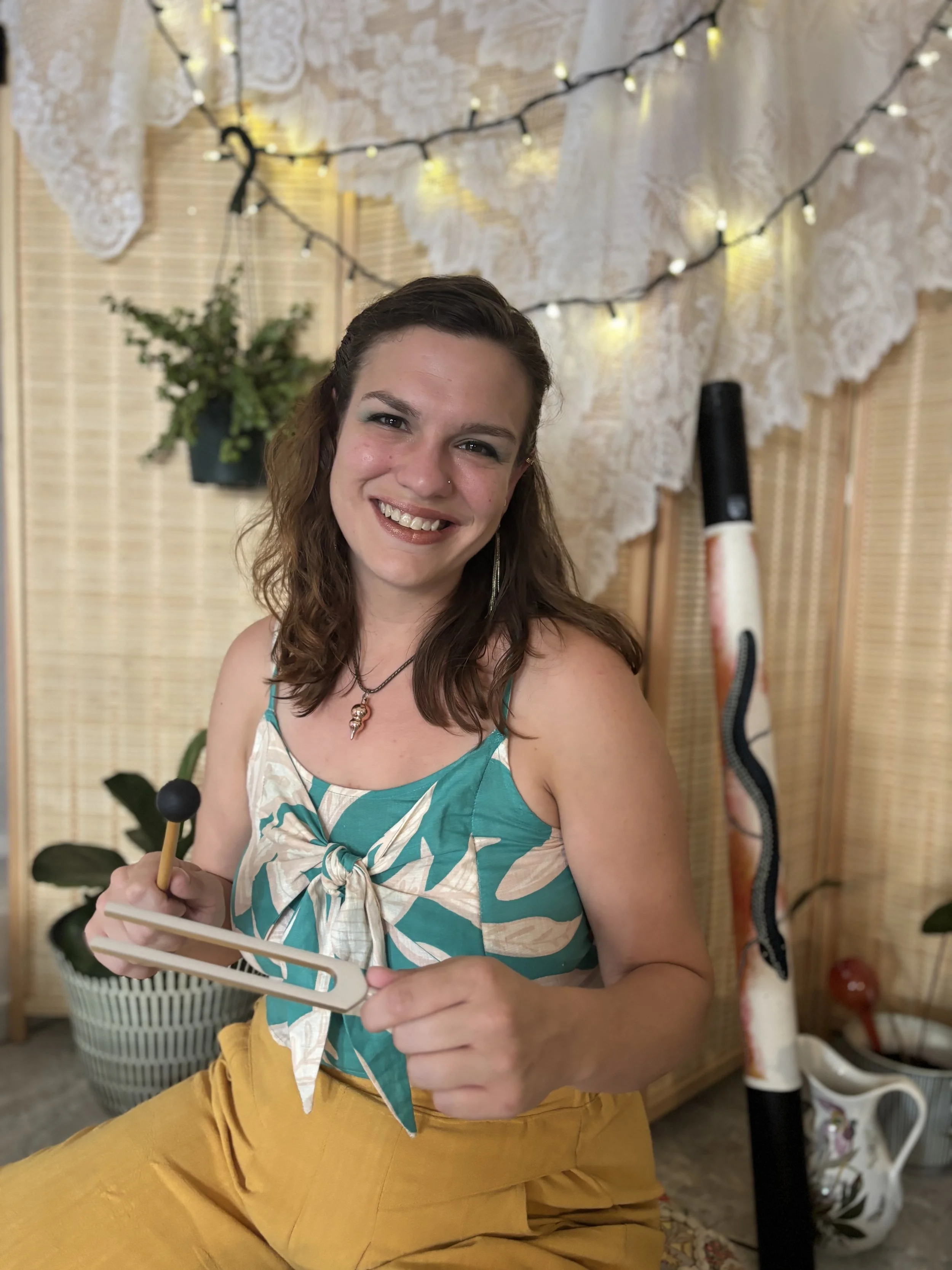 A woman sitting on a cushion on the floor, playing a set of colorful singing bowls with a mallet. She has shoulder-length brown hair, is smiling, and is wearing a blue and white patterned top with yellow pants. The background features lace curtains, potted plants, and a wave-shaped instrument standing upright.