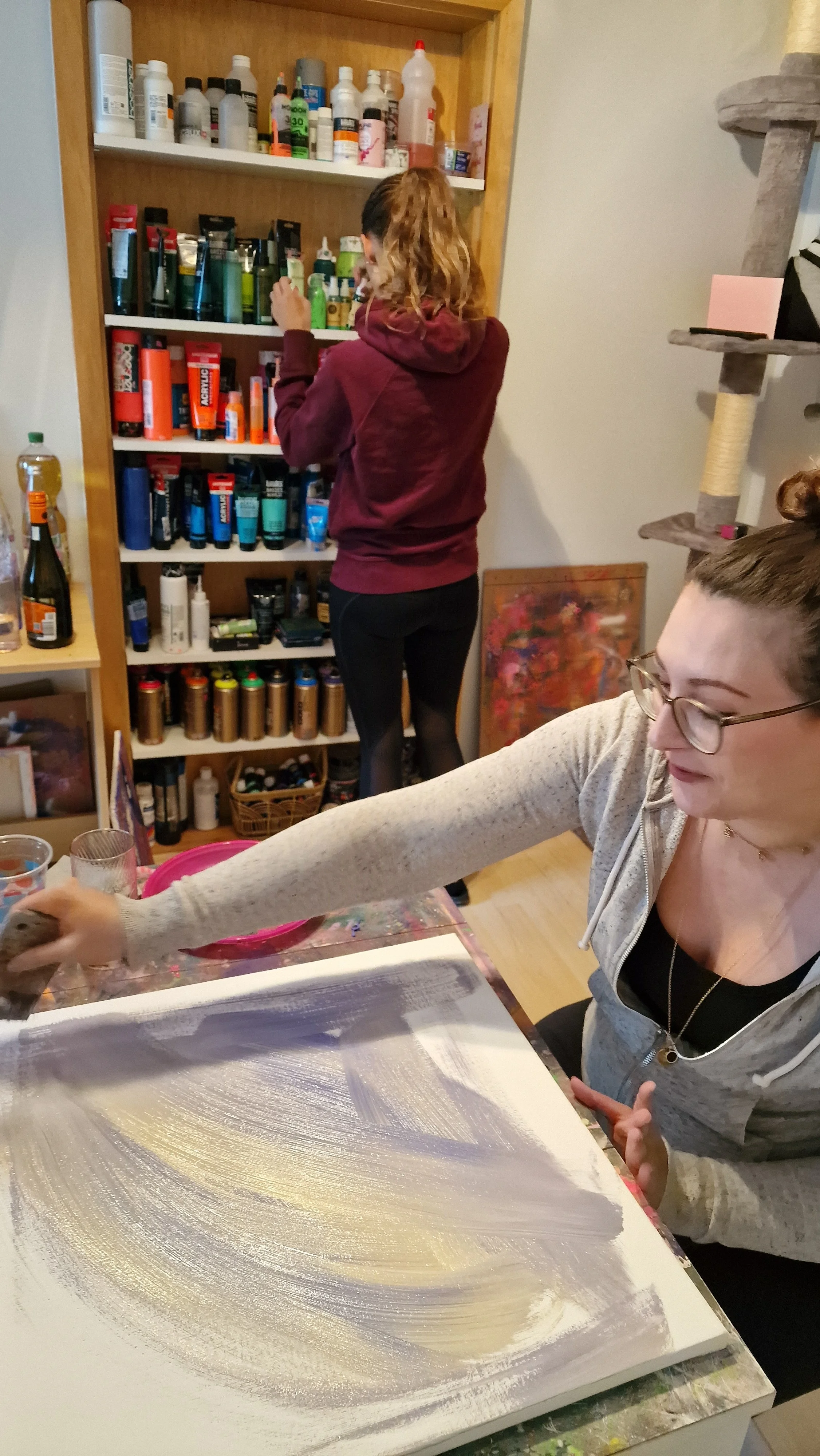 Zwei Frauen beim Malen in einem Raum, eine steht vor einem Regal mit Farbe und die andere arbeitet an einer Leinwand mit Gold- und Silberfarben.