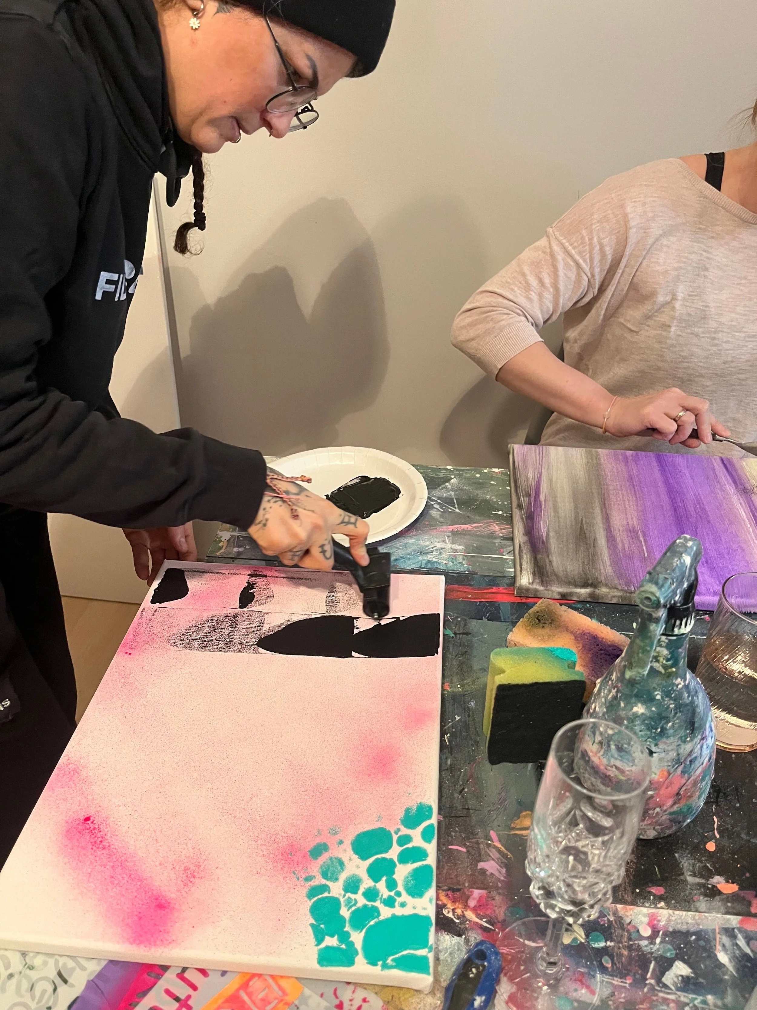 Zwei Frauen malen abstrakte Kunstwerke mit Sprühfarbe und Pinsel auf Leinwände in einem Atelier.