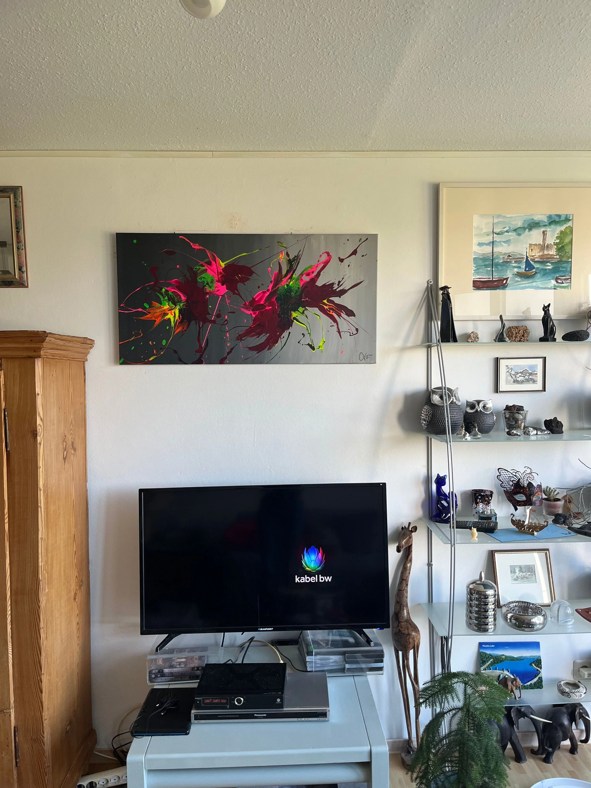 Wohnzimmerwand mit einem Flachbildfernseher, darunter ein TV-Tisch, links ein hölzerner Schrank, darüber ein abstraktes, buntes Gemälde. Rechts an der Wand befinden sich Regale mit verschiedenen Dekorationen wie Keramikeulen, eine Giraffenfigur und Pflanzen. An der Wand sind auch andere Bilderrahmen mit Kunstwerken zu sehen.