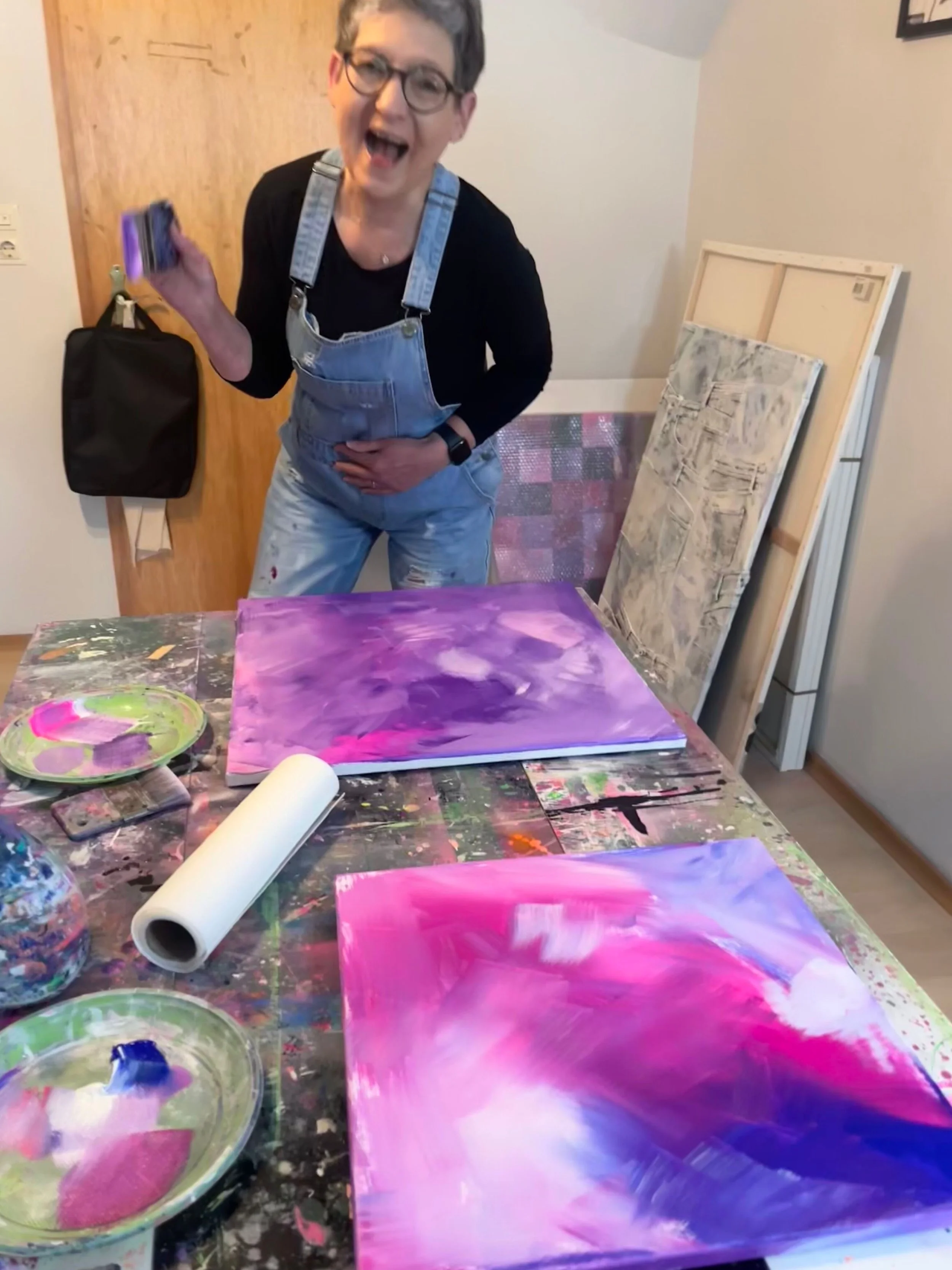 Eine Frau steht lachend in einem Kunststudio vor einem Tisch mit zwei abstrakten, violett-pinken Gemälden und Malutensilien, neben ihnen sind weitere unvollständige Gemälde im Hintergrund.