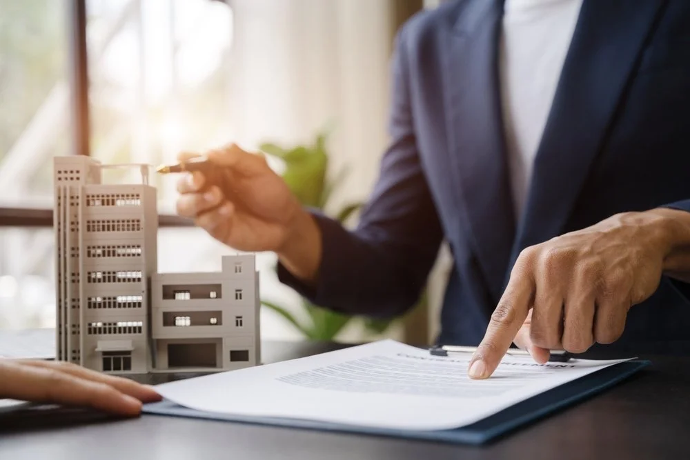 A person in a dark suit shows a miniature model of a multi-story building as another person points at a document on a clipboard on the table.