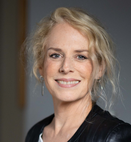Portrait of a smiling woman with blonde hair and earrings, wearing a black top, in front of a gray background.