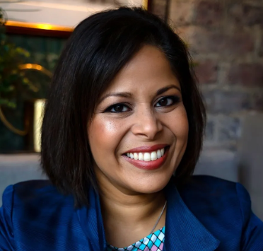 Portrait of a smiling woman with dark hair, wearing a blue blazer, in front of a brick wall.