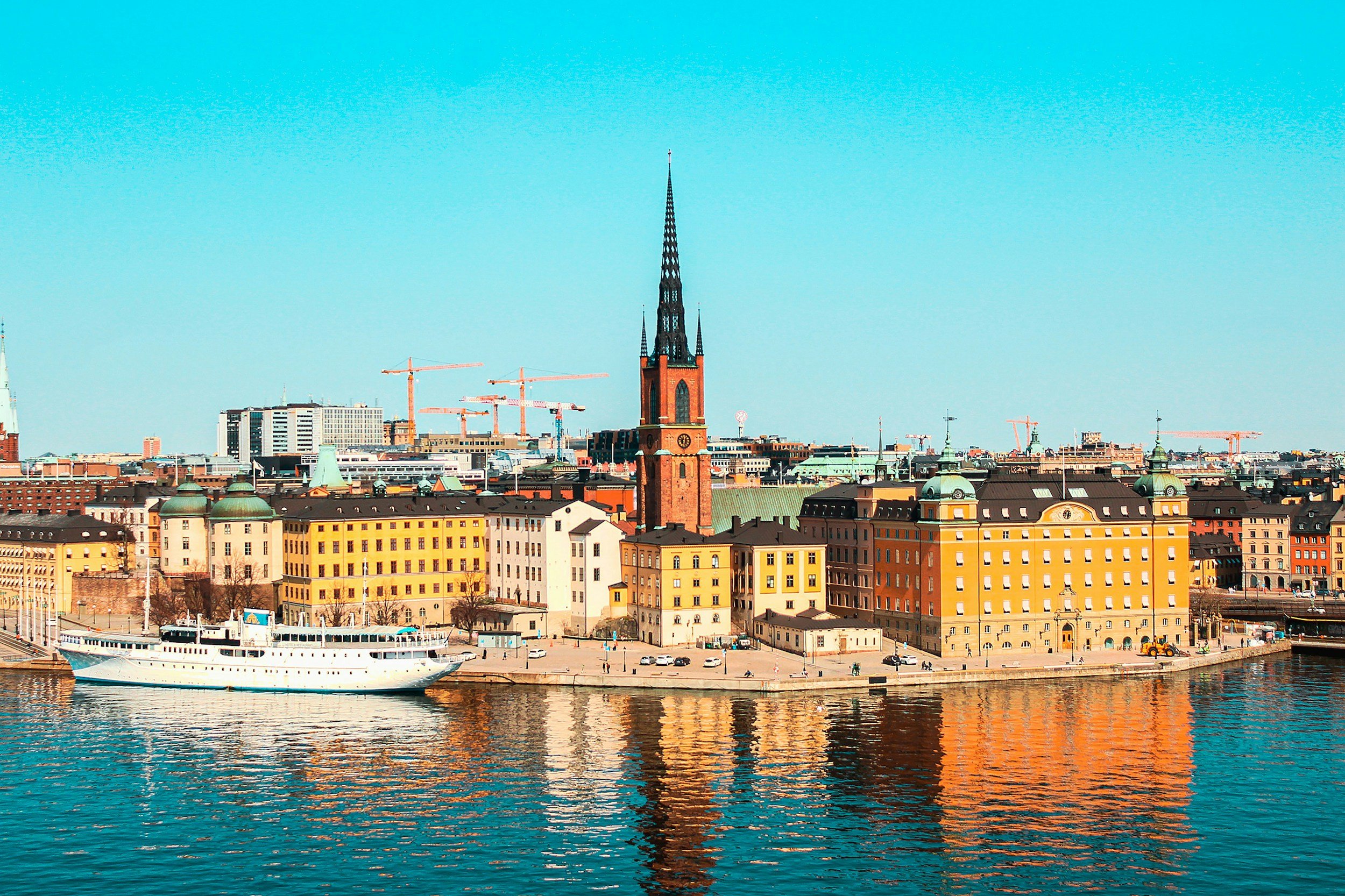 Panoramic view of a city waterfront with colorful historic buildings, a white boat on the water, and a tall church steeple against a blue sky.