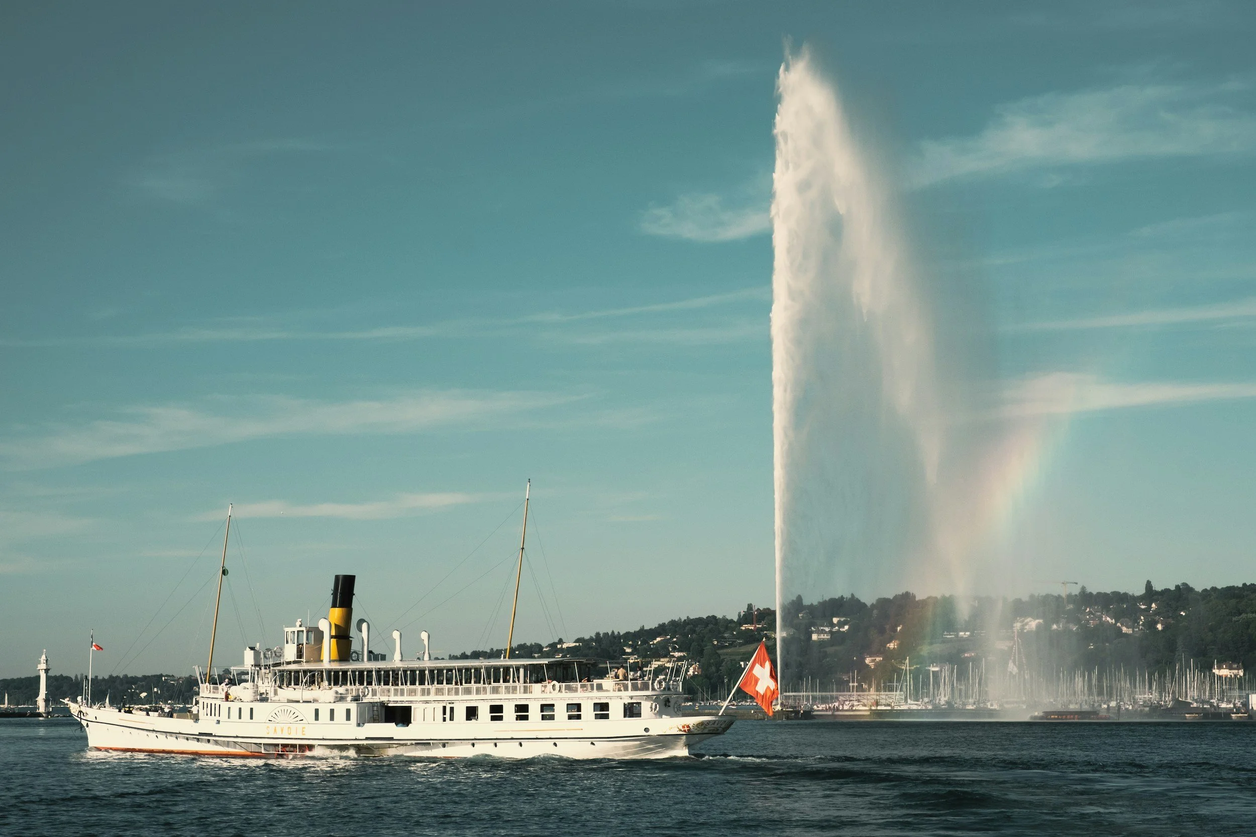 A large white boat with a black and yellow smokestack on water, flying a Swiss flag. A tall water jet erupts from the water near the boat, creating a rainbow in the mist. Land with trees and houses are visible in the background.