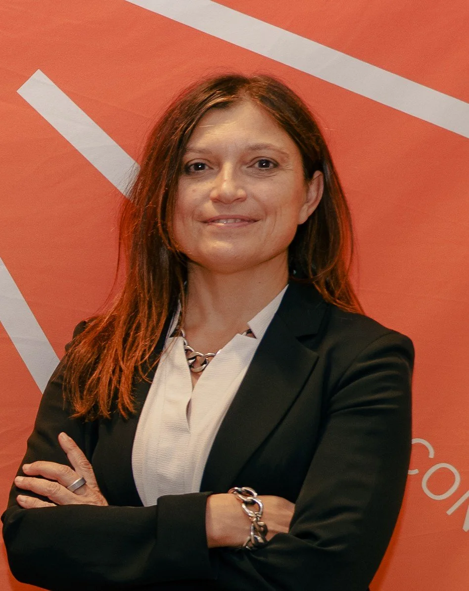 A woman with brown hair wearing a black blazer, white shirt, and silver jewelry, standing with arms crossed in front of an orange background with white diagonal stripes.