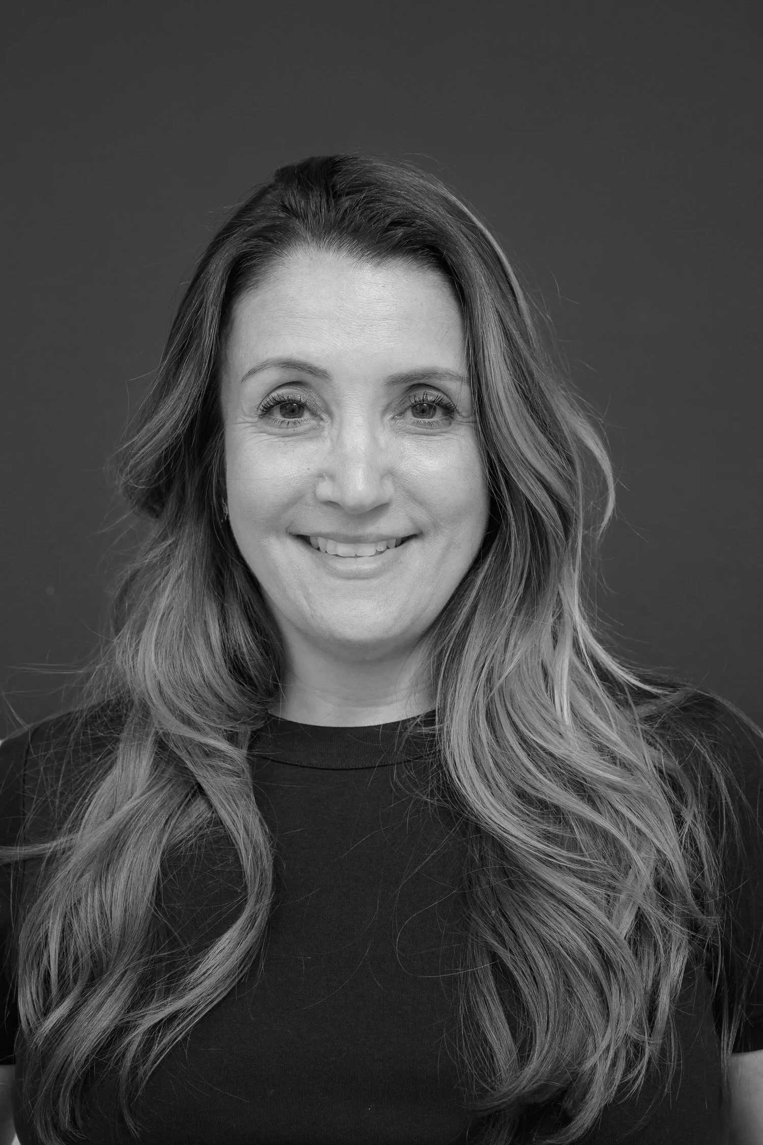 Black and white headshot of a woman with long wavy hair, smiling, wearing a dark top against a plain background.