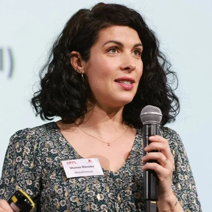 A woman with dark, curly hair speaking into a microphone at a presentation or conference, wearing a floral-patterned top and a name tag labeled 'Shanaz Diessler, SleepSensor'.