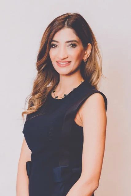 A young woman with long, wavy brown hair wearing a sleeveless dark dress, standing against a plain light-colored background, smiling at the camera.