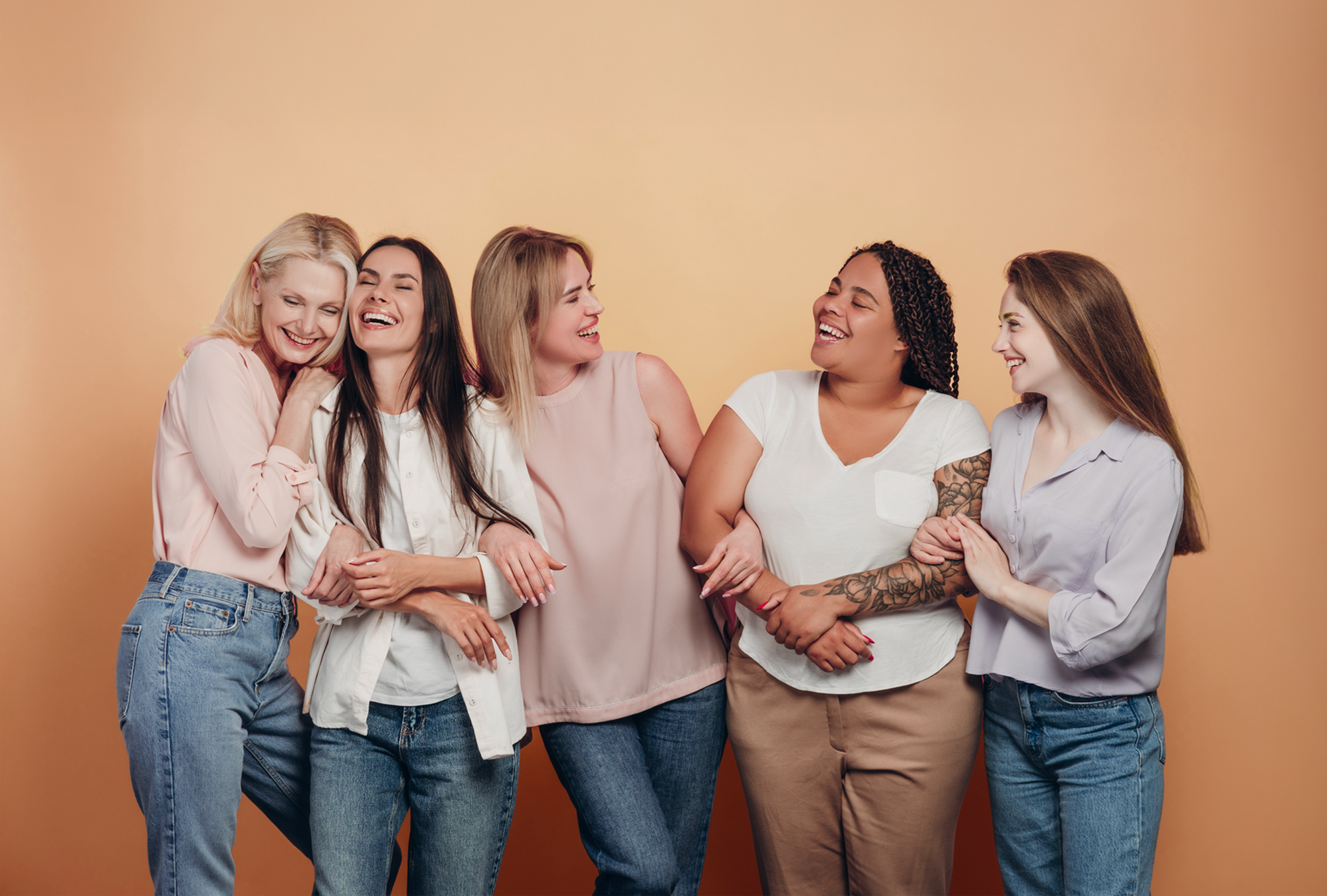 Six women standing together, laughing and smiling, against a yellow background.