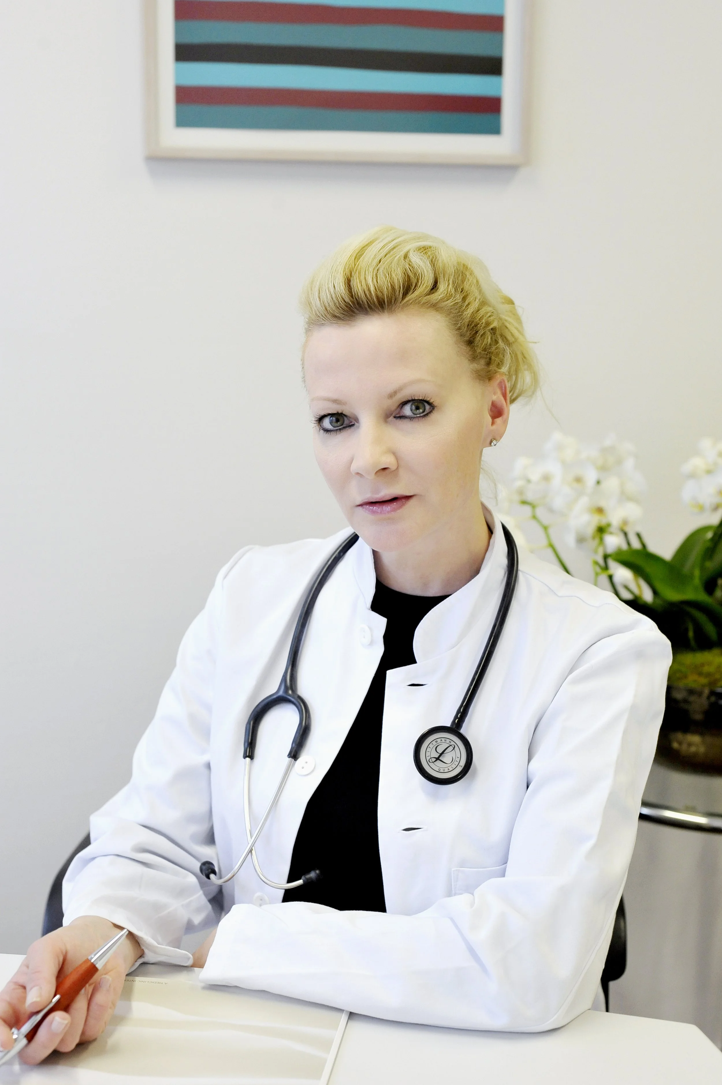A female doctor with blond hair, wearing a white lab coat and stethoscope, sitting at a desk with a pen, in a medical office setting, with white orchid flowers and a framed picture on the wall behind her.