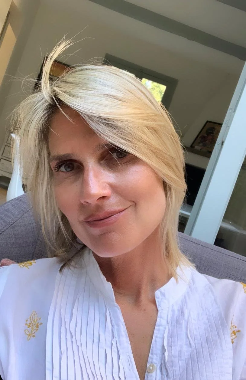 A woman with blonde hair and light skin taking a selfie indoors, sitting on a gray couch, with room windows and artwork in the background.