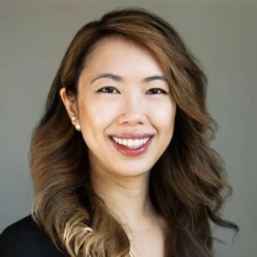 Portrait of a smiling woman with long, wavy hair wearing pearl earrings and a black top against a gray background.
