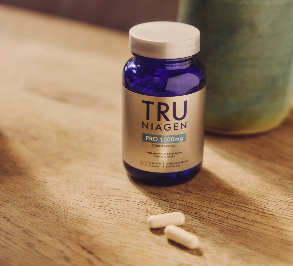 A bottle of TRU Niagen PRO 1000MG dietary supplement capsules on a wooden surface with two capsules in front of it.
