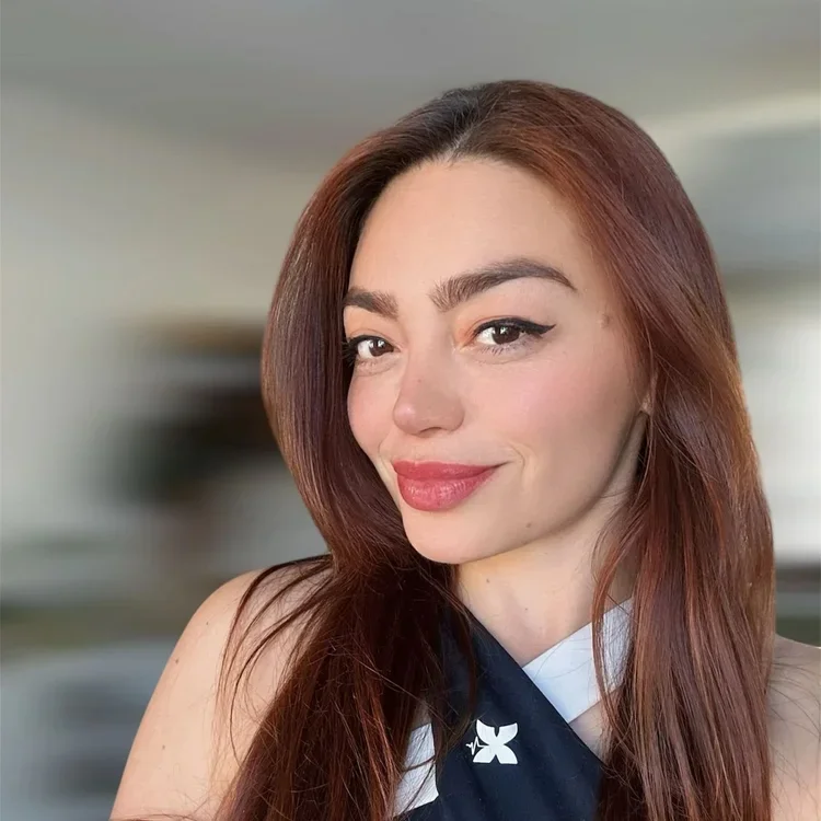 Close-up of a young woman with long, reddish-brown hair, smiling slightly with her lips closed, wearing winged eyeliner and a sleeveless top with a scarf.