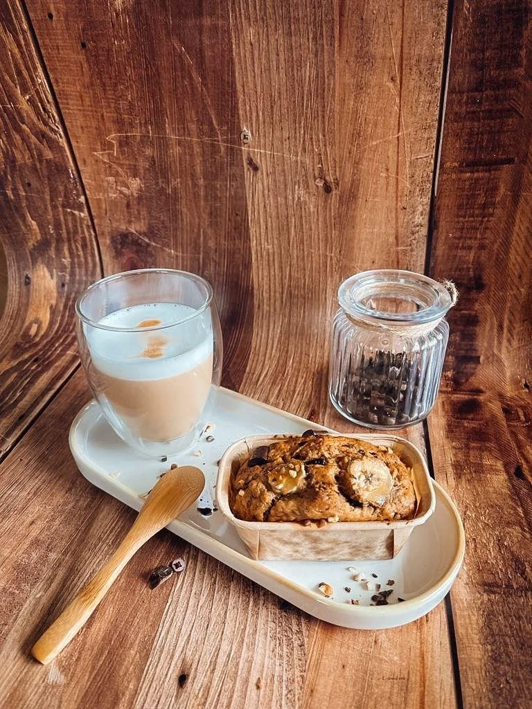 A wooden table with a half-full glass of iced coffee topped with foam, a small jar of coffee beans, a wooden spoon, and a baked good that looks like a cookie or bread with nuts and chocolate chips on a white tray.