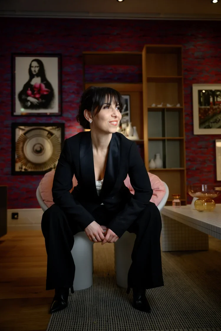 A woman in a black suit sitting on a white chair in a modern living room with red textured wall, framed artwork, and wooden shelving.
